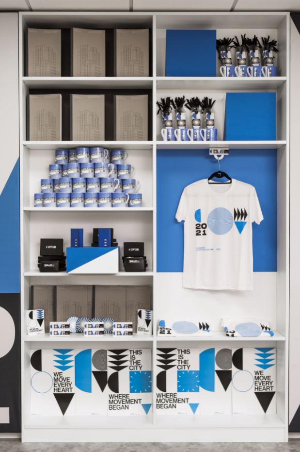 Image: White shelves stocked with City of Culture merchandise including mugs, t-shirts, wallets, magnets and coasters. Photo by Lewis Copson.
